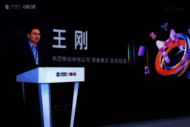 王刚在台上发表讲话，身后大屏展示咪咕云游戏战略布局图，包含技术架构与生态合作模式
